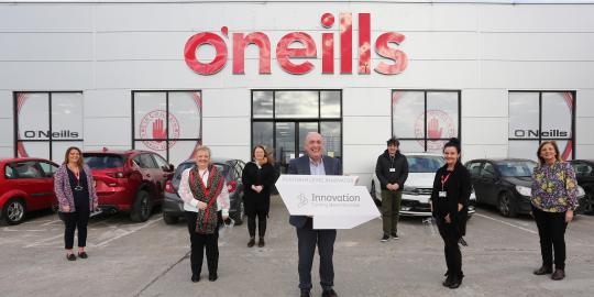 O'Neills staff holding Platinum level innovator badge outside O'Neills factory in Strabane, Co. Tyrone
